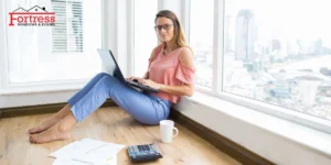 Woman working on laptop by a window with natural light, papers and calculator on floor, Fortress Windows and Doors branding Woman working on laptop by a window with natural light, papers and calculator on floor, Fortress Windows and Doors branding